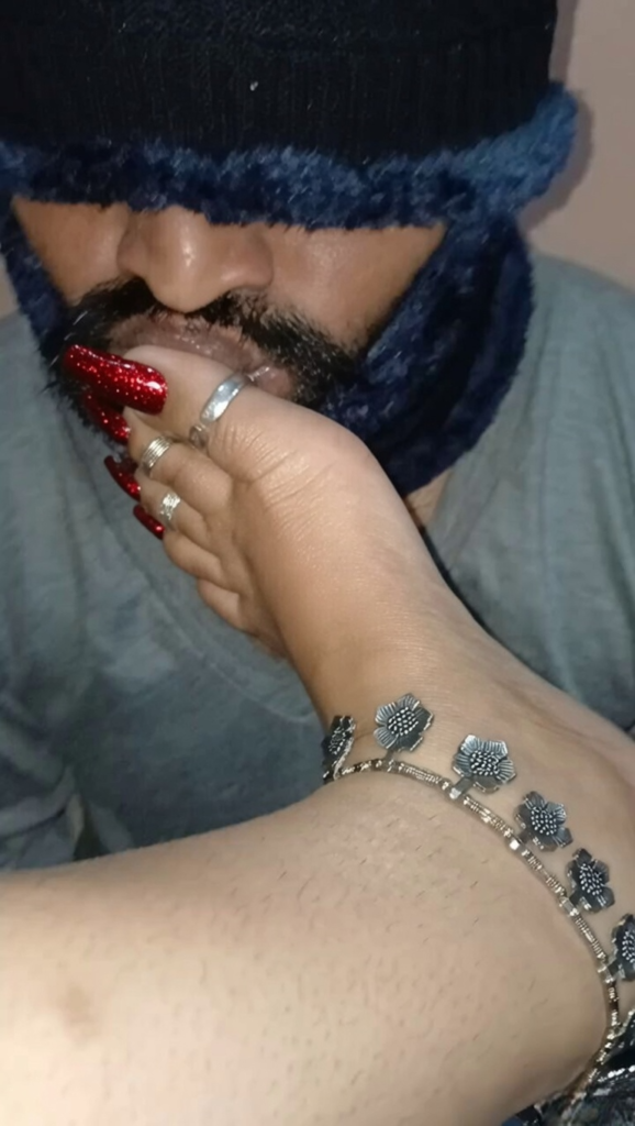 Elite Dubai BDSM Mistress session showcasing authentic foot worship as a submissive male client kisses her foot adorned with a silver floral anklet and rings, while long red glitter stiletto fingernails gently cup his chin.
