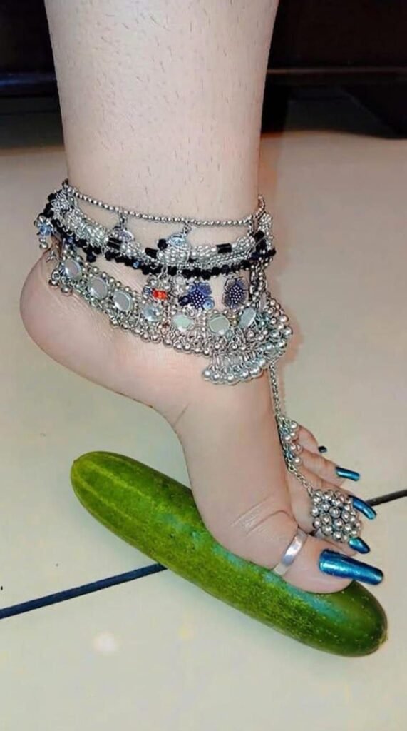 Close-up of New York City BDSM Mistress Anu's foot with blue metallic nail polish, wearing a heavy traditional silver payal (anklet) with a toe ring while stepping on a green cucumber.