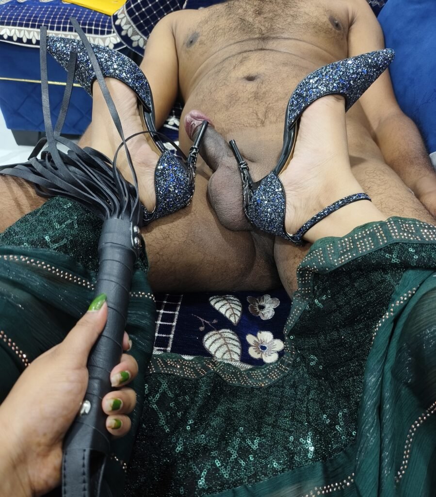 Pattaya Bdsm Mistress Anu holding a black leather flogger whip while resting her blue glitter stiletto heels on a submissive male's groin.