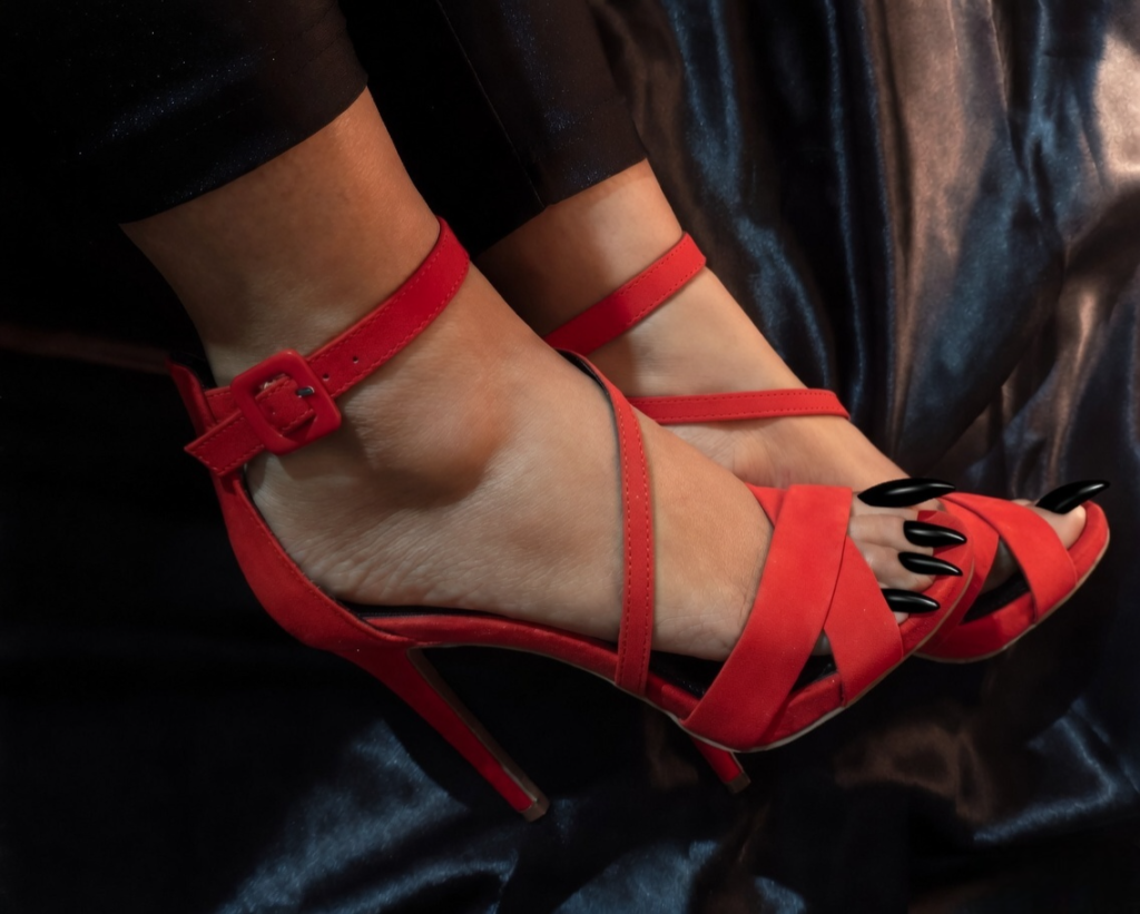 Close-up of Penang BDSM Mistress Anu wearing elegant red strappy high-heeled sandals with long black manicured toenails against a dark silky background.
