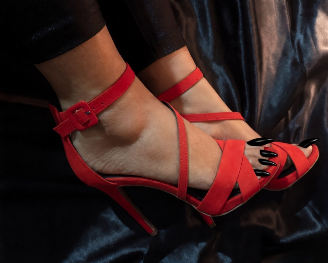 Close-up of Penang BDSM Mistress Anu wearing elegant red strappy high-heeled sandals with long black manicured toenails against a dark silky background.